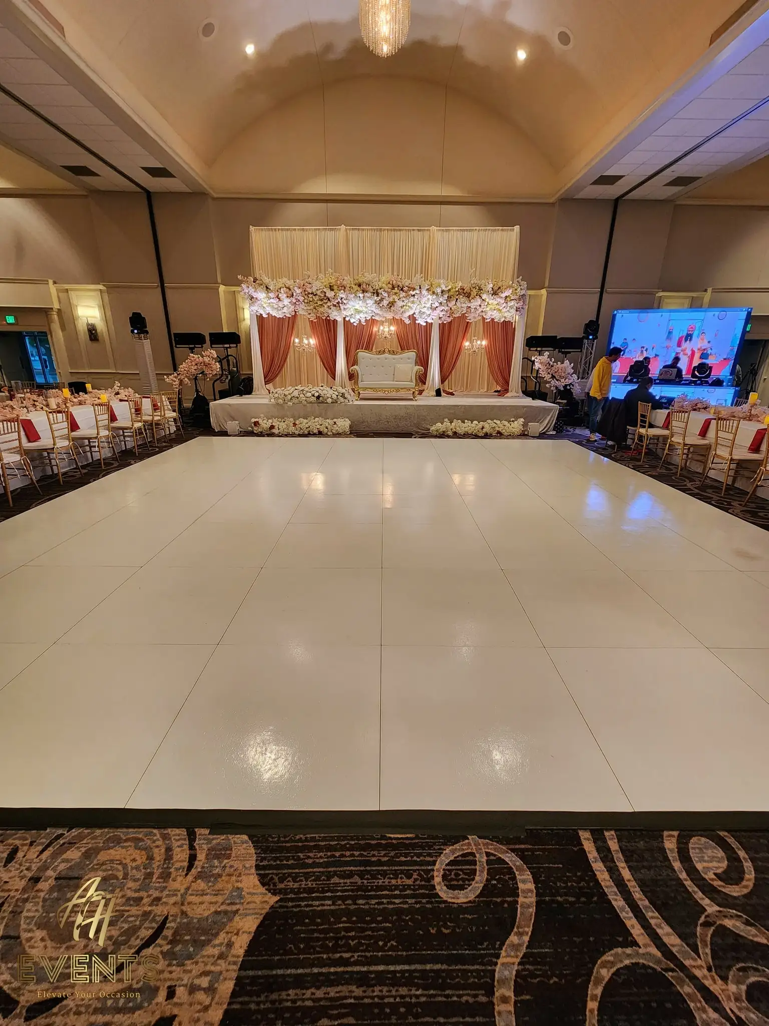 White dance floor facing a cream-and-gold draped stage with floral garlands and a tufted sofa, an AV screen to one side