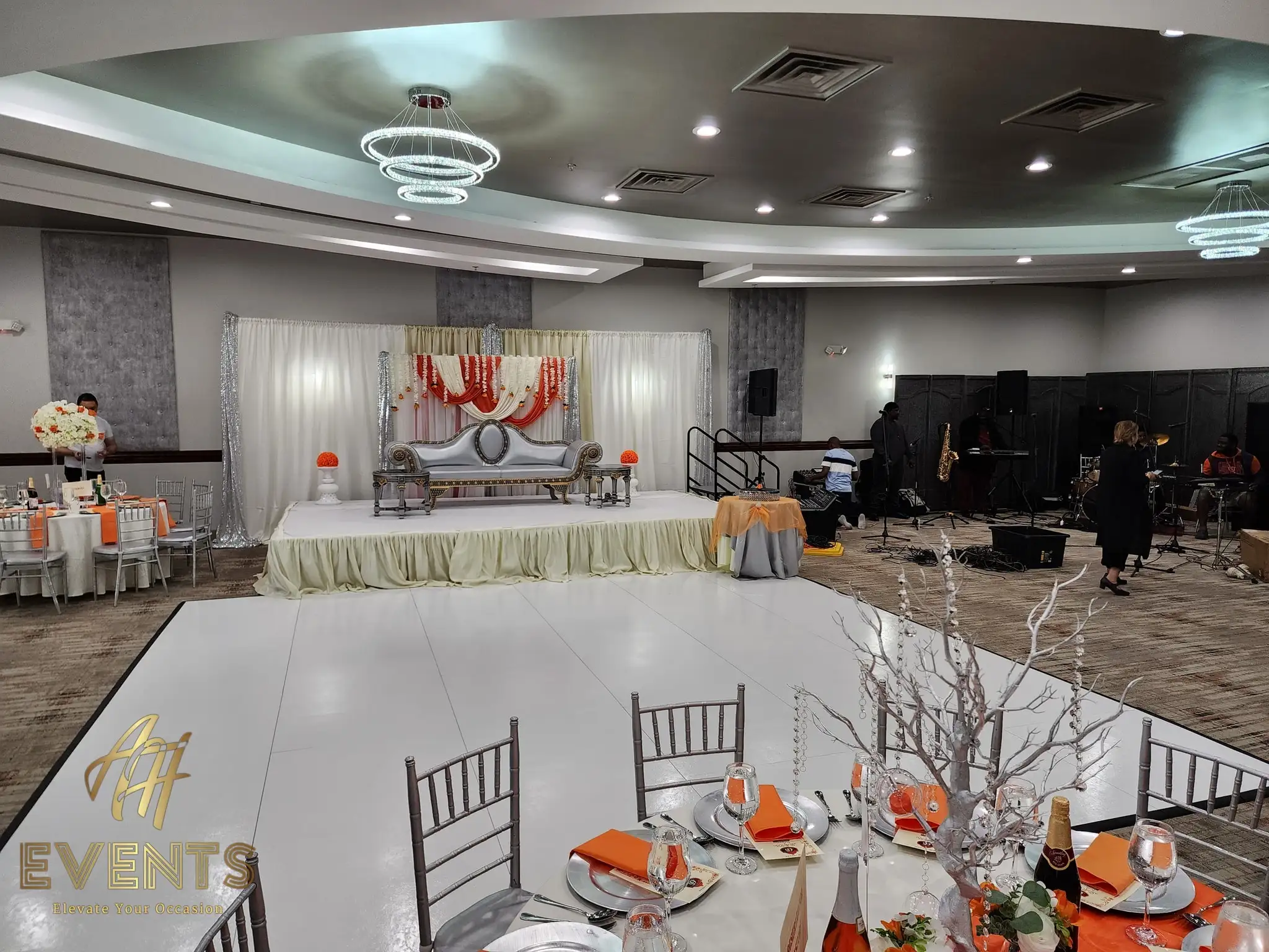 White dance floor before a cream-and-gold stage with a silver sofa, orange table linens and a live band setup