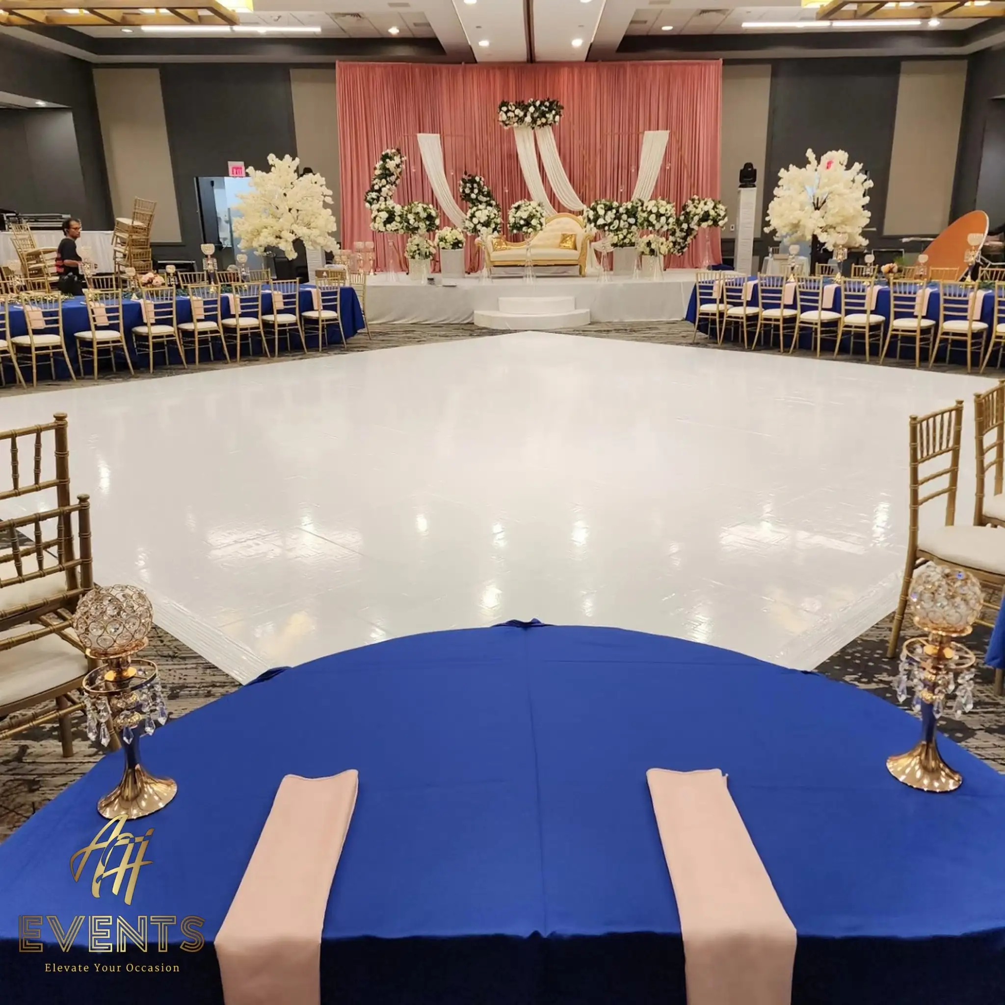 White dance floor edged by royal-blue tables, facing a blush-draped stage with white florals