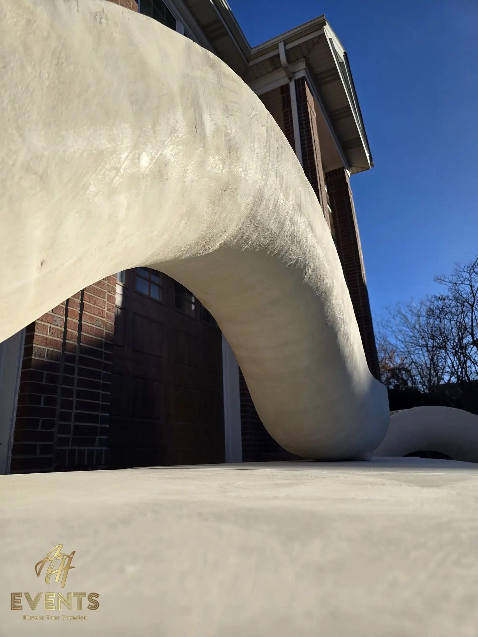 Close low-angle of a single sculpted white serpentine arch form, a brick house behind