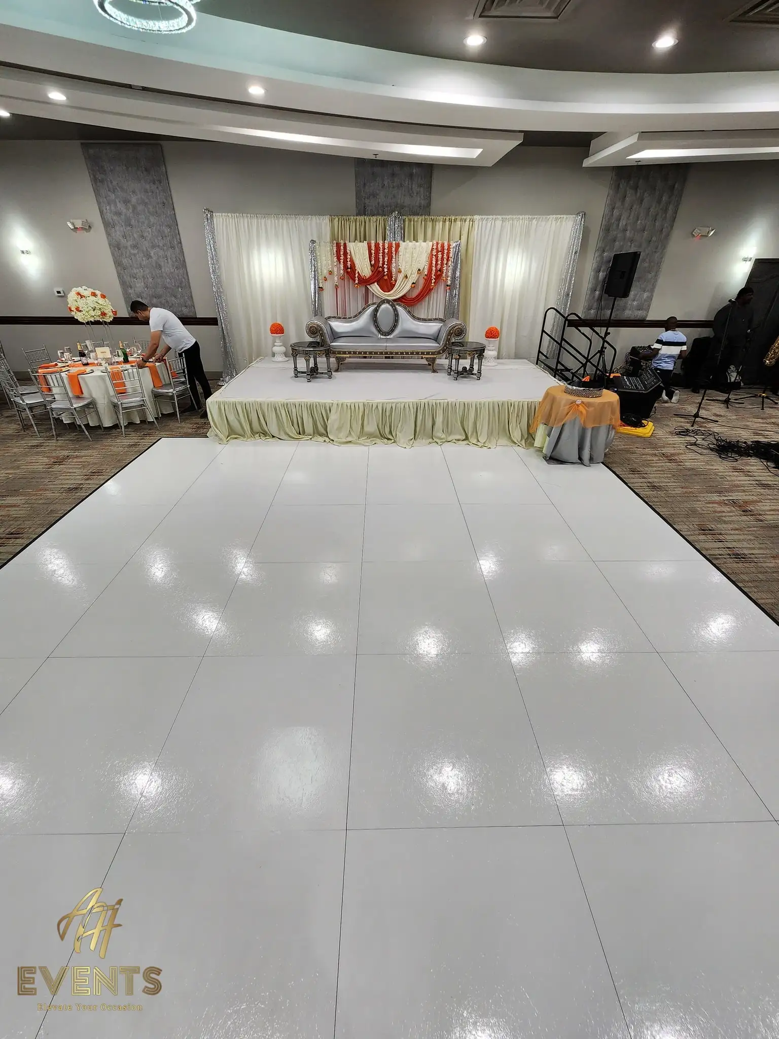 White dance floor during setup, facing a cream stage with a silver sofa and a red-and-cream floral swag