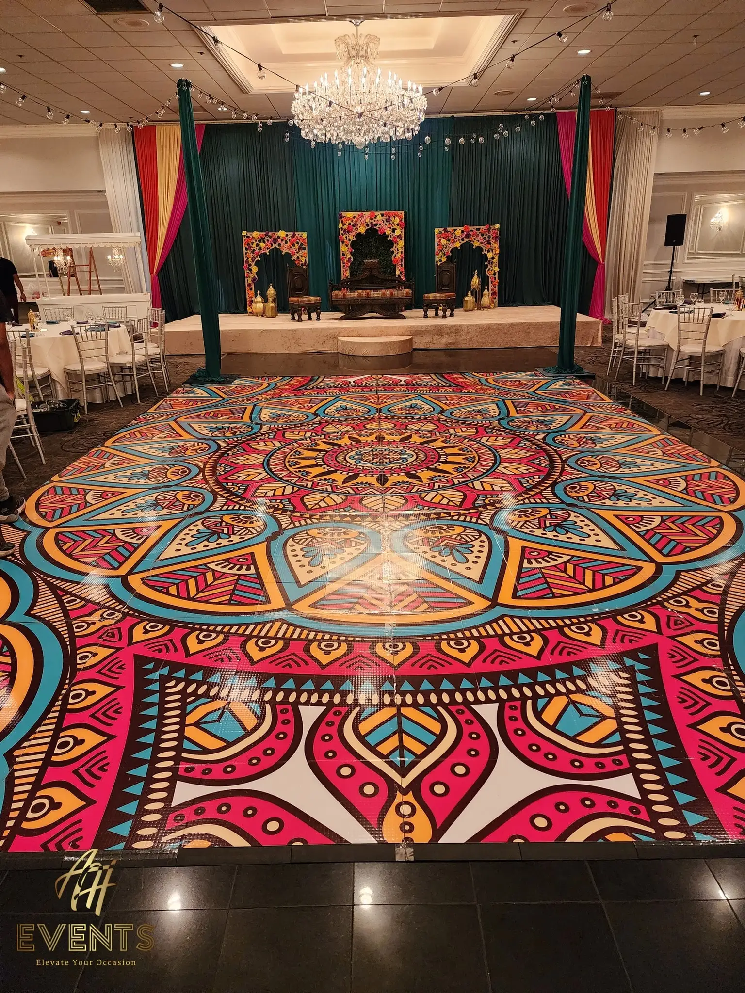 Mandala dance floor beneath a chandelier, facing a teal-draped stage with a carved throne