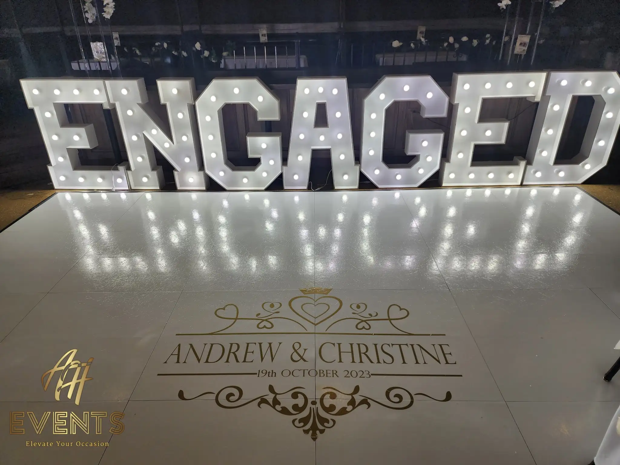 Light-up 'ENGAGED' marquee letters on a white floor printed with an 'Andrew & Christine' gold monogram decal