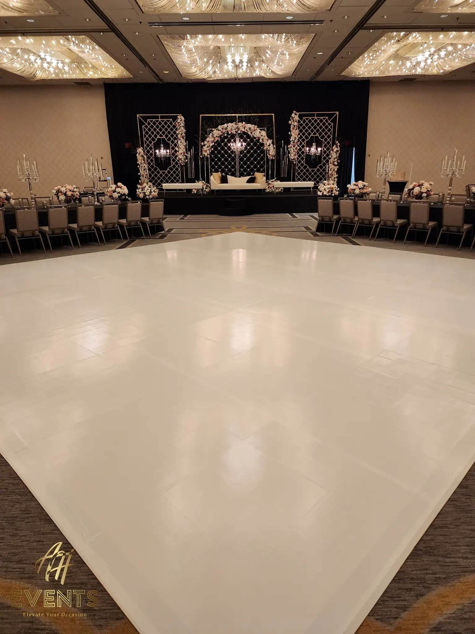 Glossy white dance floor before a black stage with a floral mandap and crystal chandeliers, silver chairs around