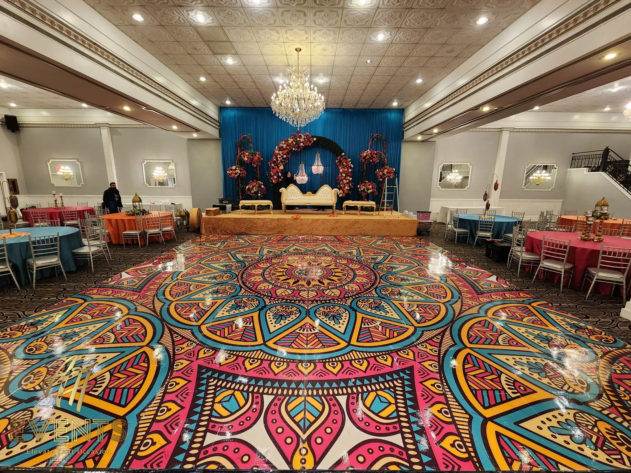 Large mandala floor facing a blue-draped stage with a floral arch and sofa, colorful table linens around