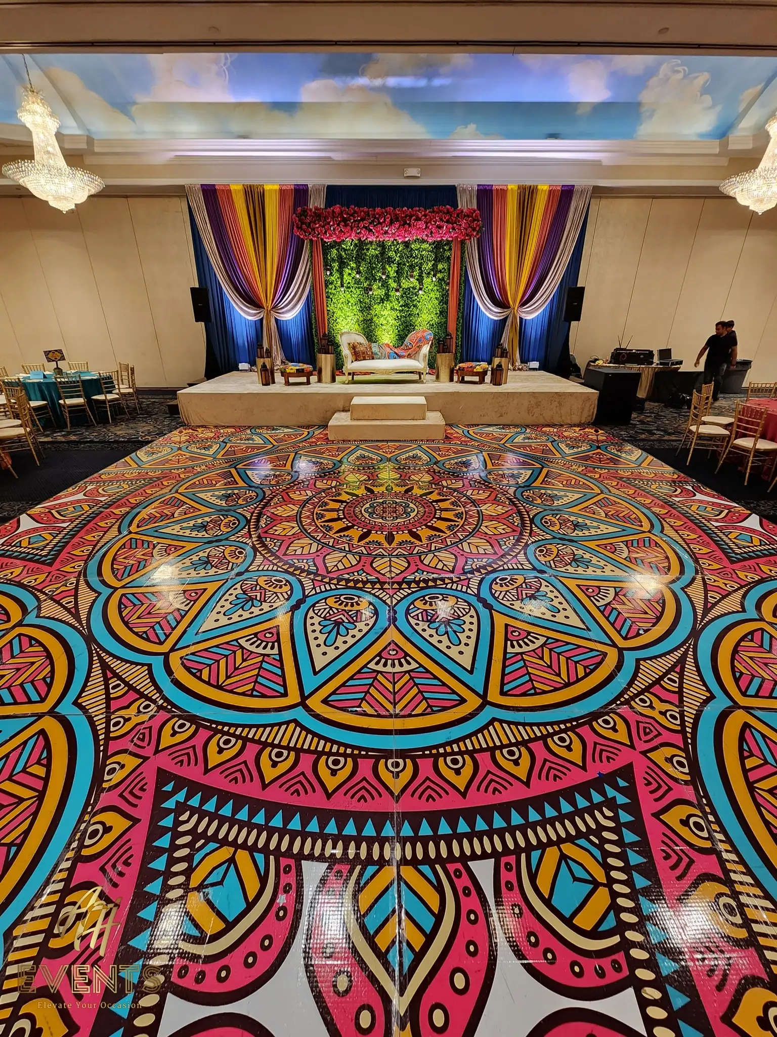 Mandala dance floor under a cloud-painted ceiling, facing a blue-draped stage with a greenery arch