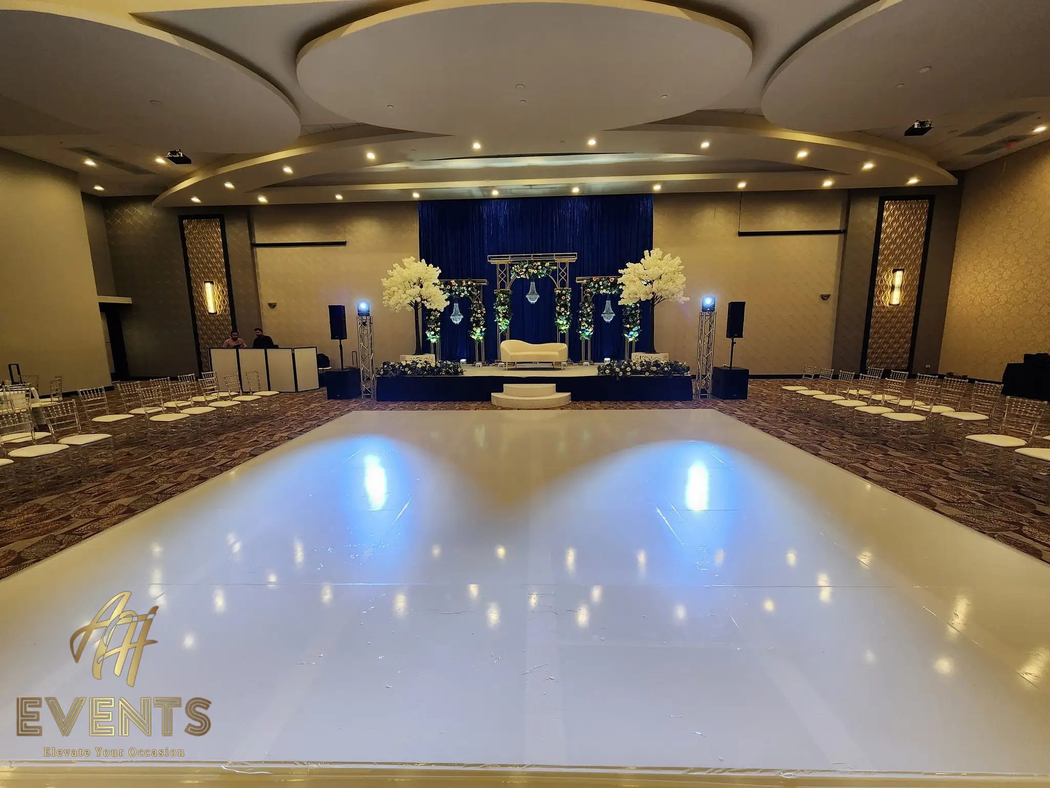 Glossy white dance floor facing a navy stage with white blossom trees and a floral mandap