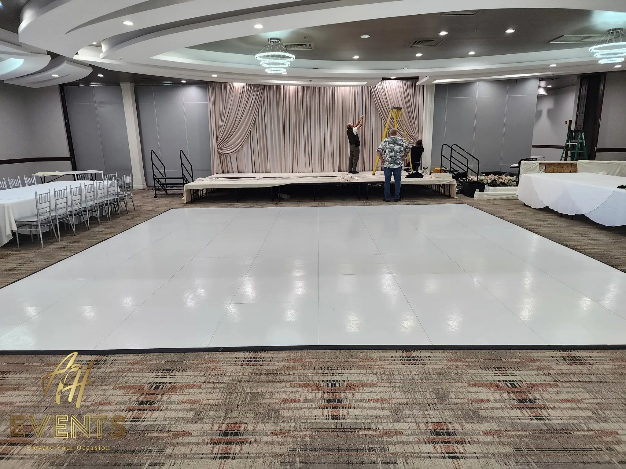 White dance floor with a taupe-draped stage under construction during event setup