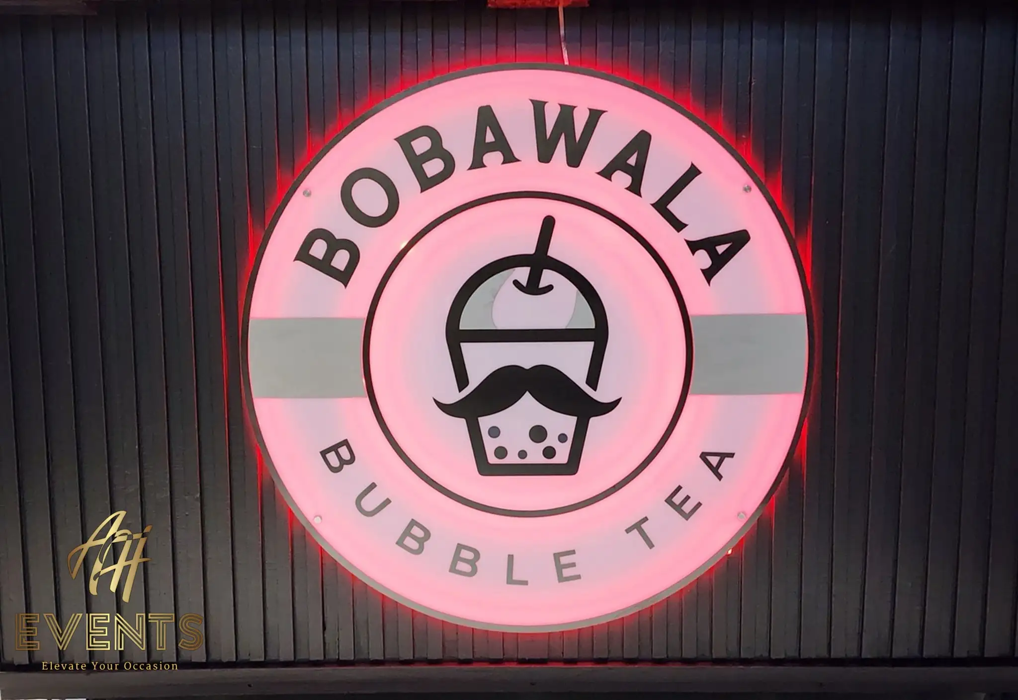Close-up of the Bobawala Bubble Tea logo roundel illuminated in pink-red on the black slatted front