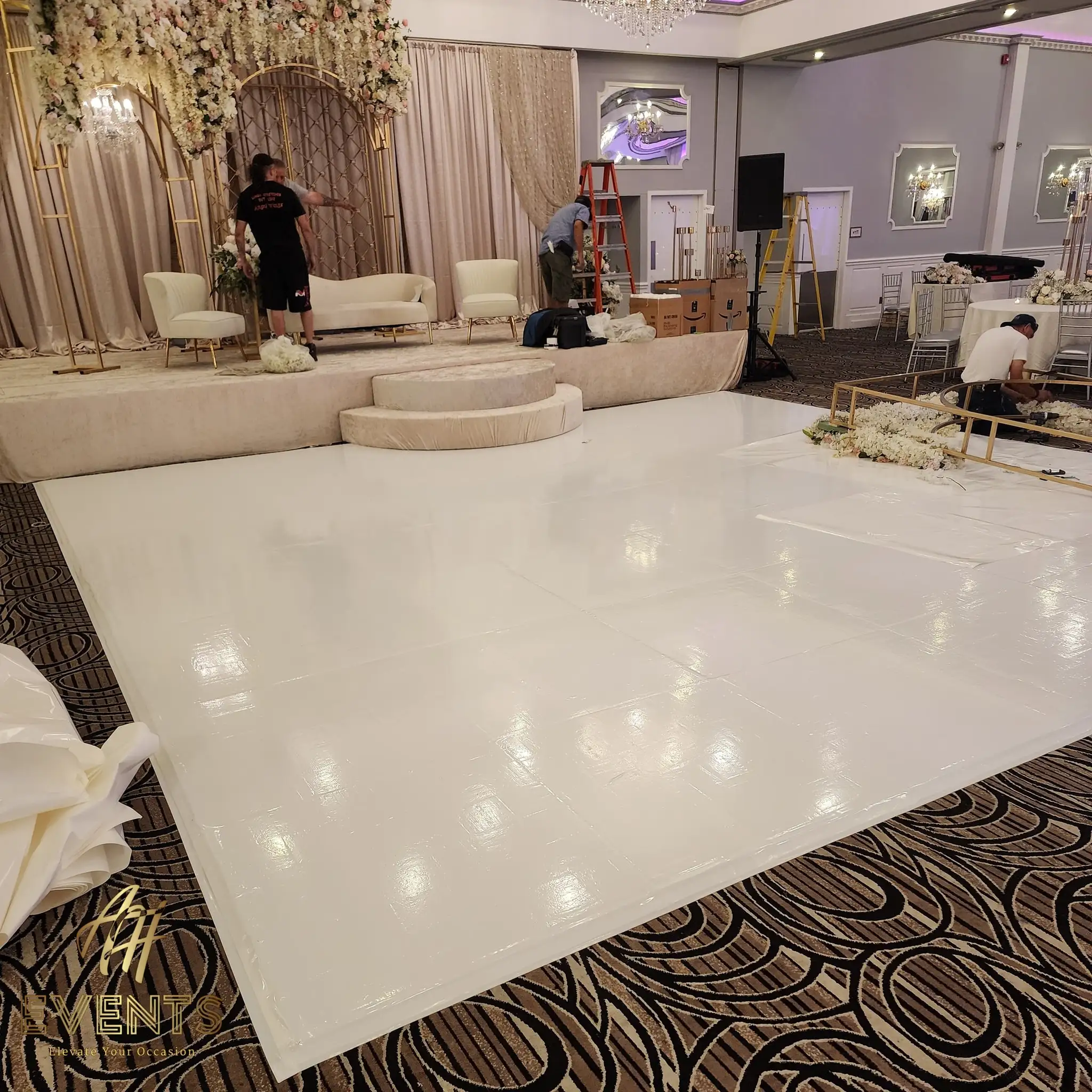 White vinyl dance floor being laid during setup, installers working by a blush stage