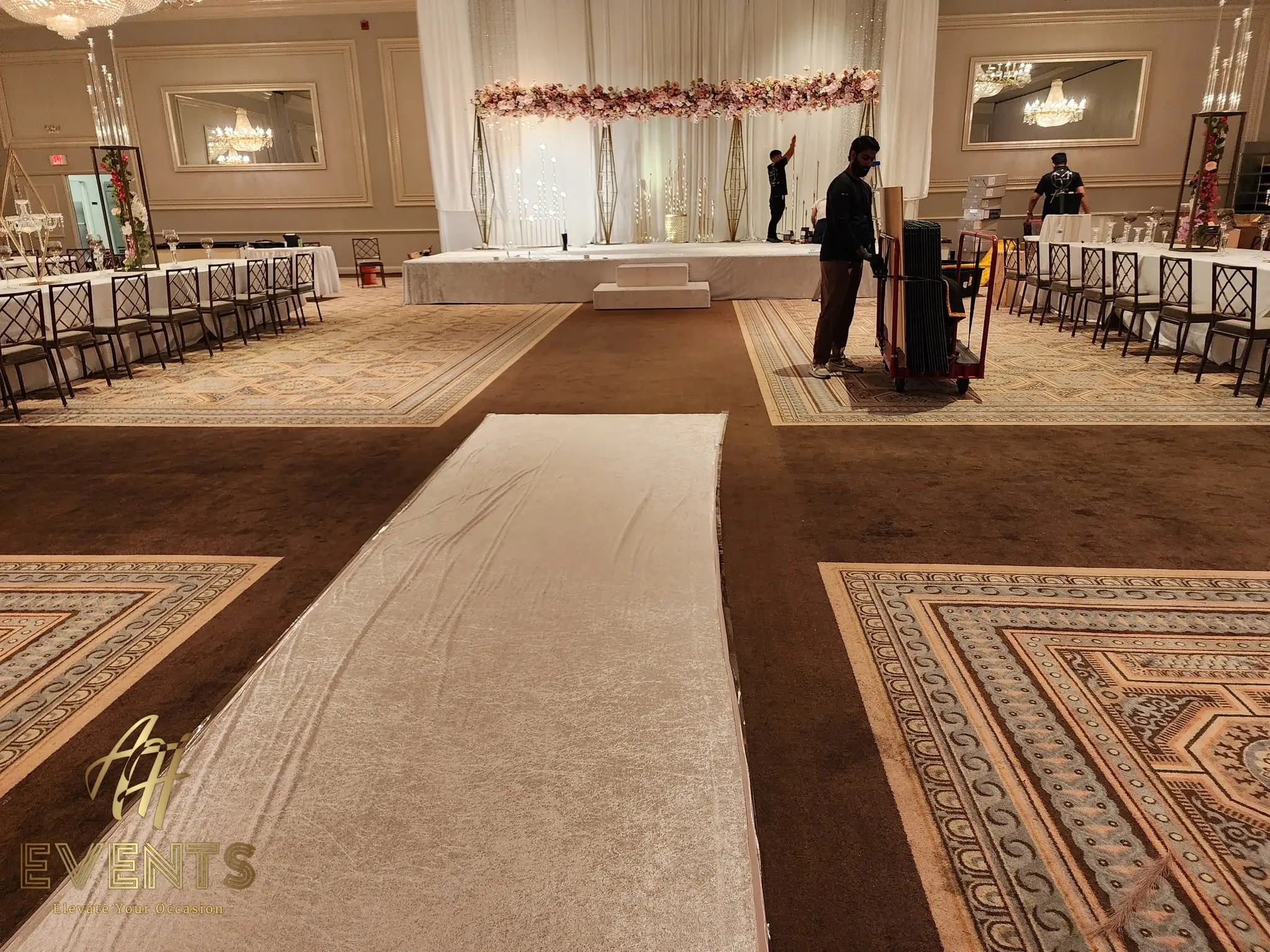 White dance floor with a white aisle runner being laid toward a blush floral stage, brown patterned carpet around