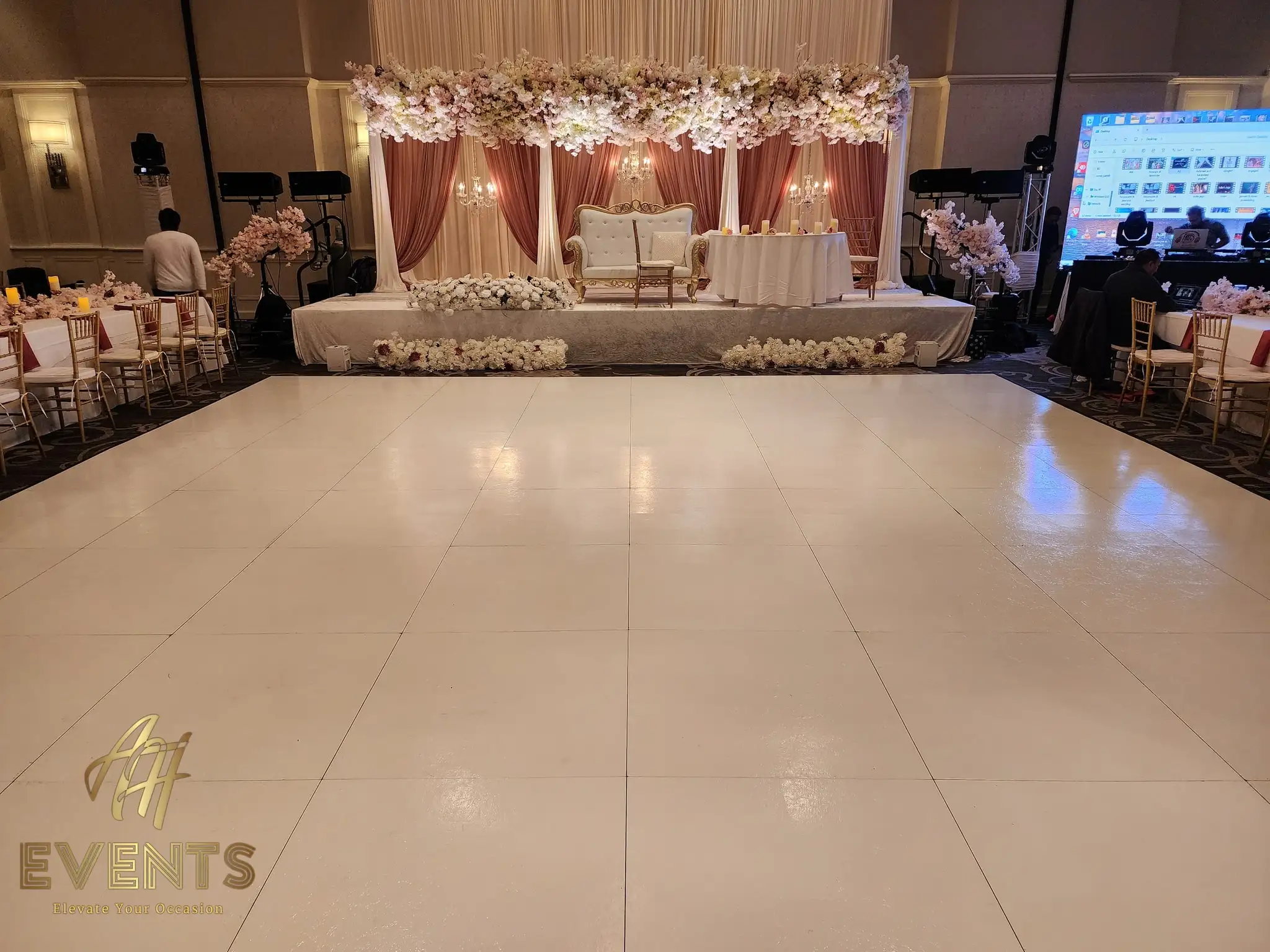 White dance floor before a mauve-draped stage with a gold loveseat under blush blossoms, an AV screen to the side