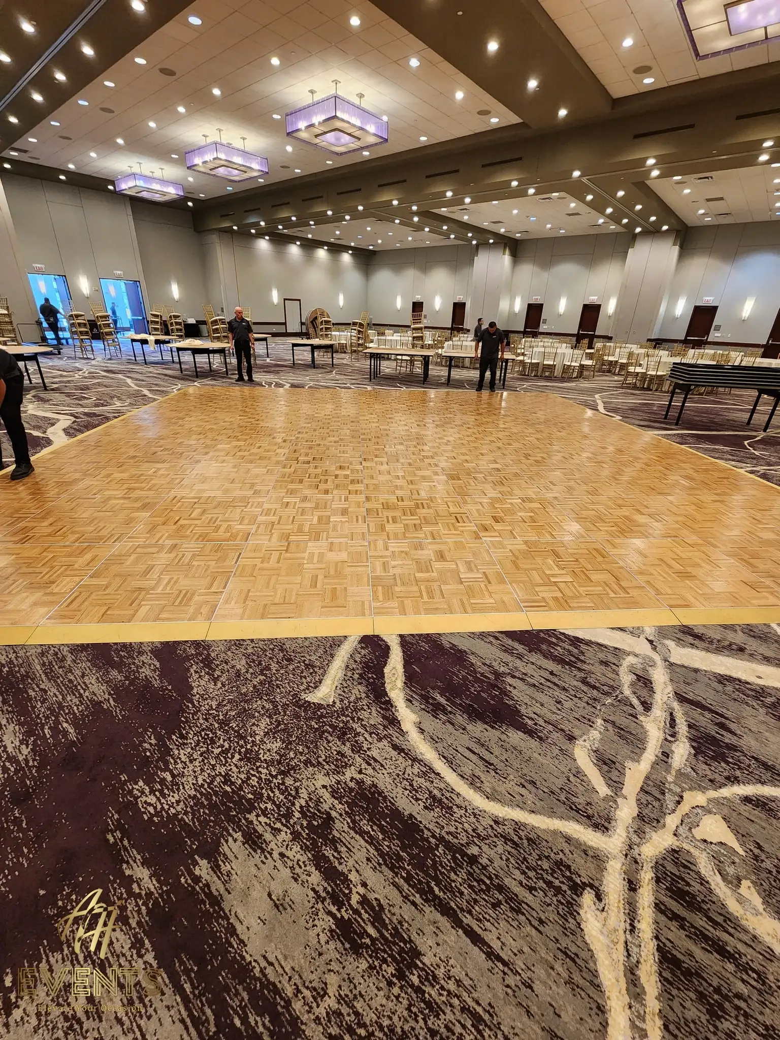 Parquet wood dance floor in a large ballroom during setup, round tables and gold chairs behind