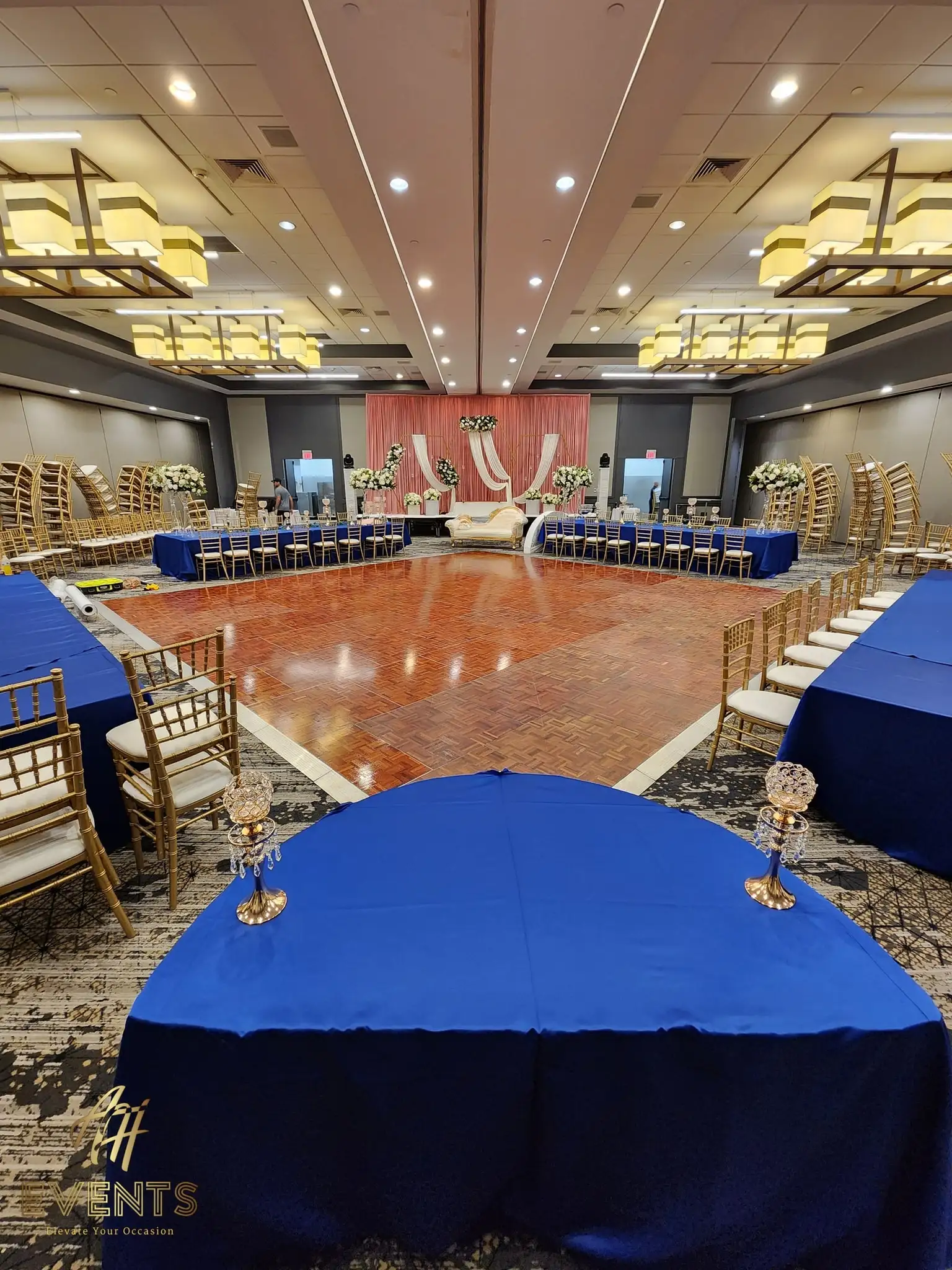 Parquet wood dance floor framed by royal-blue tables, facing a blush stage with a gold sofa
