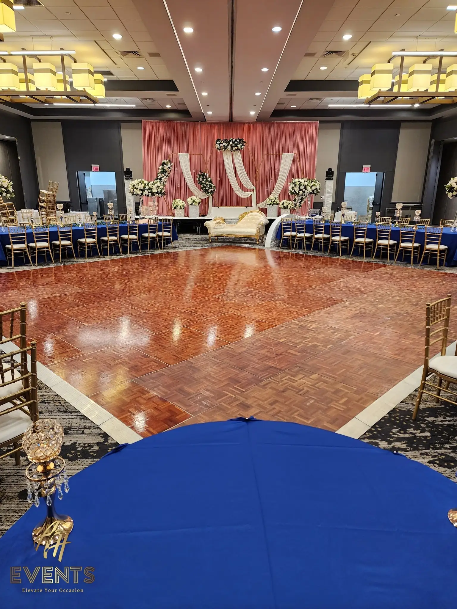Parquet wood dance floor with royal-blue tables, facing a blush-draped stage and gold sofa