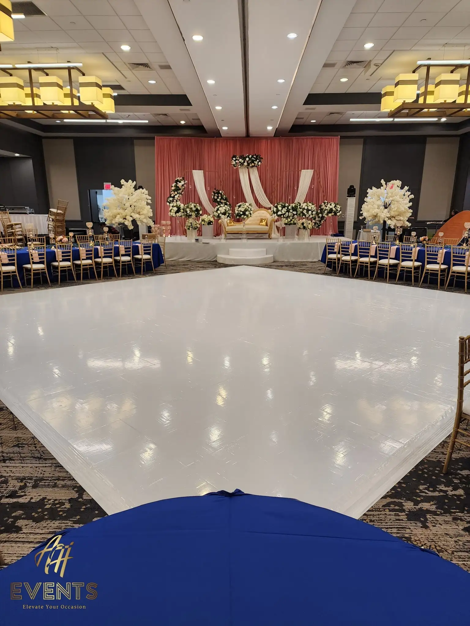 Glossy white dance floor facing a coral-draped stage with white blossom trees and a gold sofa, blue-linen tables around