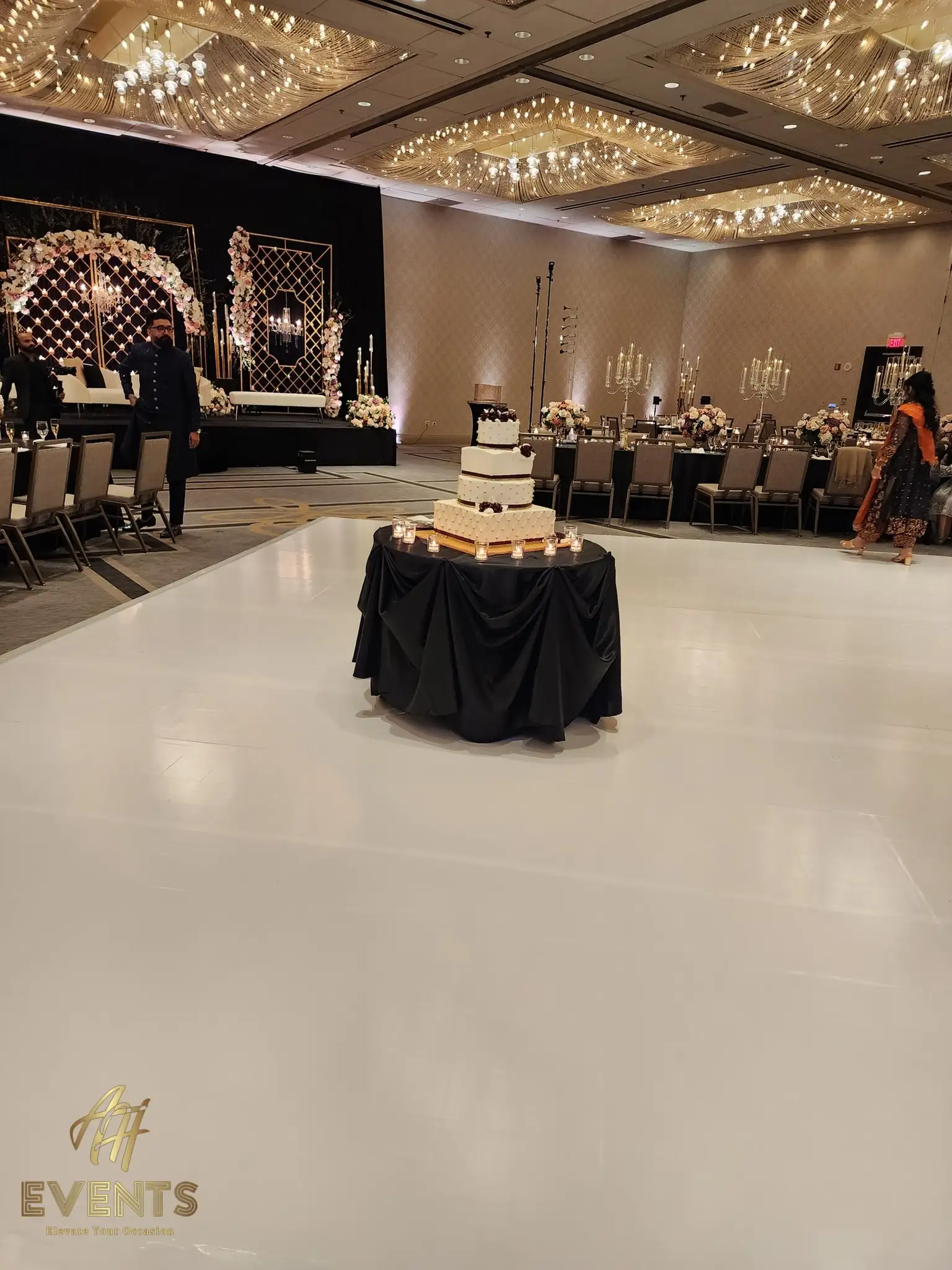 Glossy white dance floor with a black-draped cake table holding a tiered cake, the black mandap stage behind