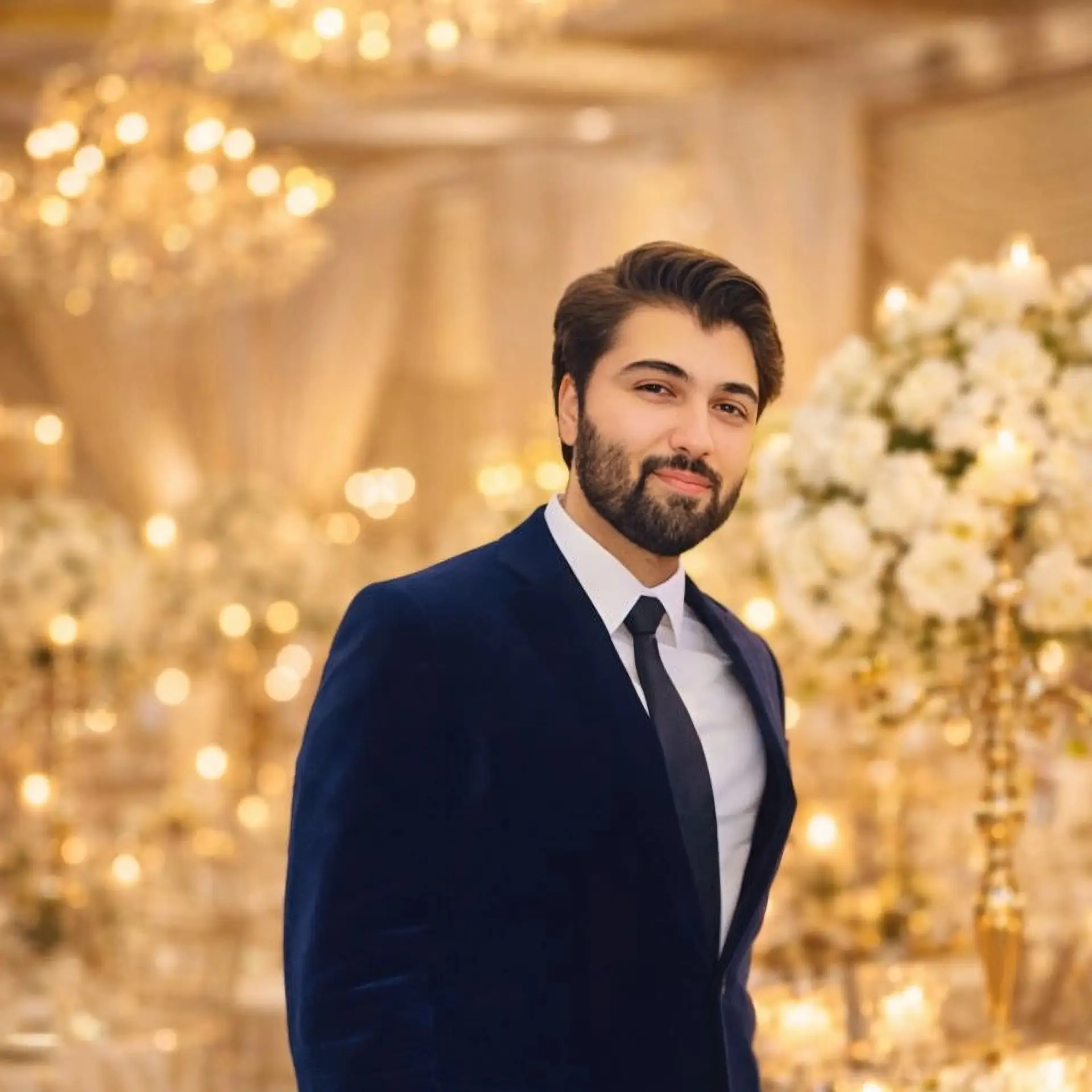 Man with dark hair and a trimmed beard wearing a plum blazer and black shirt, posed indoors and looking slightly to the side.