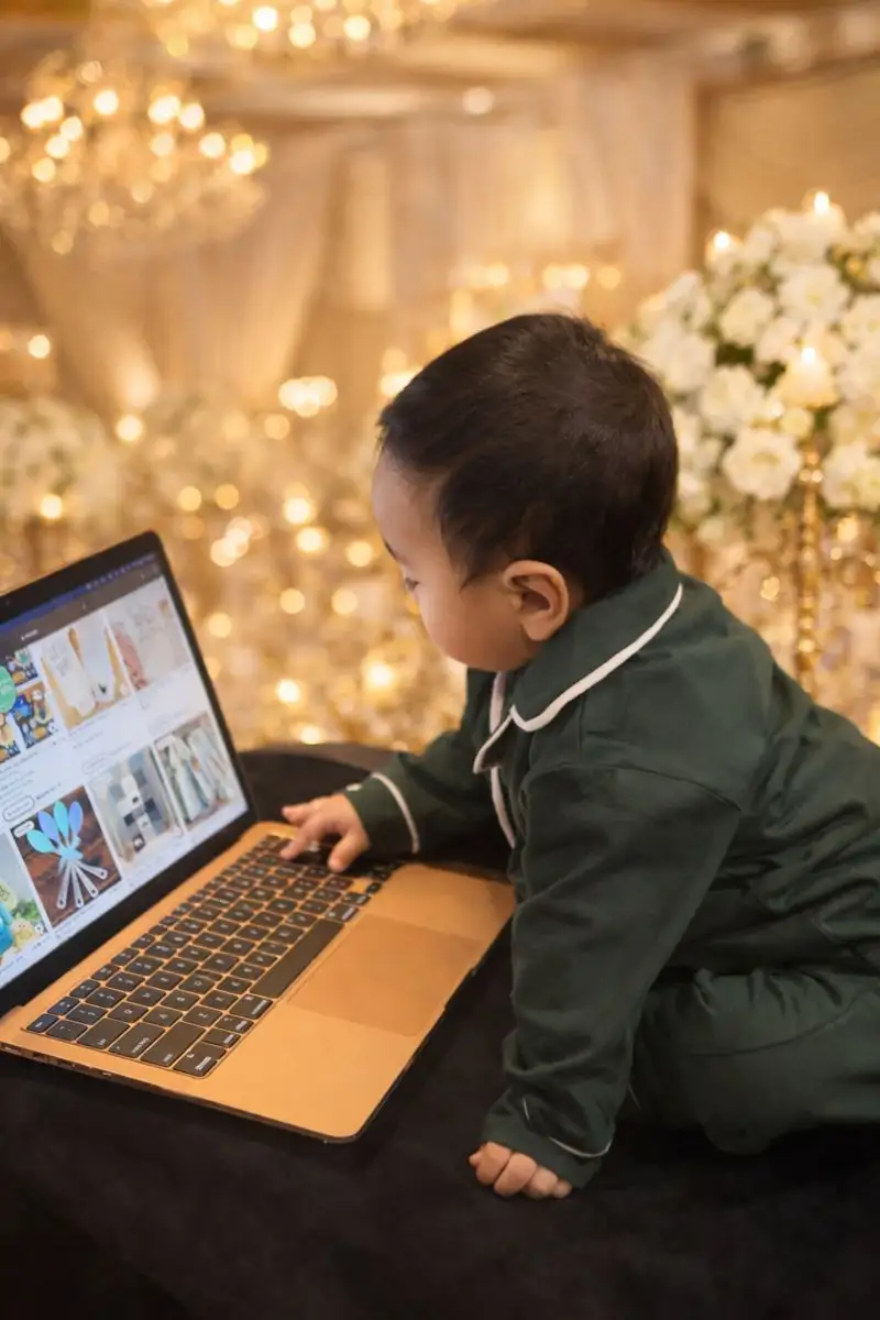 Toddler in green pajamas leaning on a couch while touching the keyboard of an open laptop.